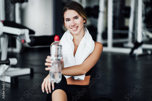 Attractive smiling girl on the floor in the middle of the gym holds water in her hands and looks at the camera