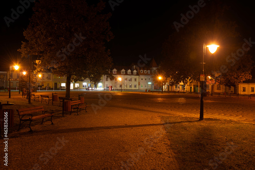 Centralny plac w małym miasteczku nocą, oświetlony światłem latarni ulicznych.