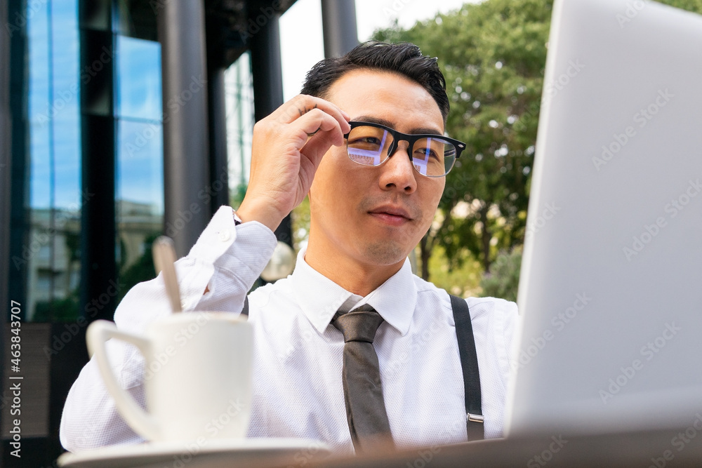 Stylish Asian businessman putting on eyewear against laptop in cafe Stylish Asian businessman putting on eyewear against laptop in cafe
