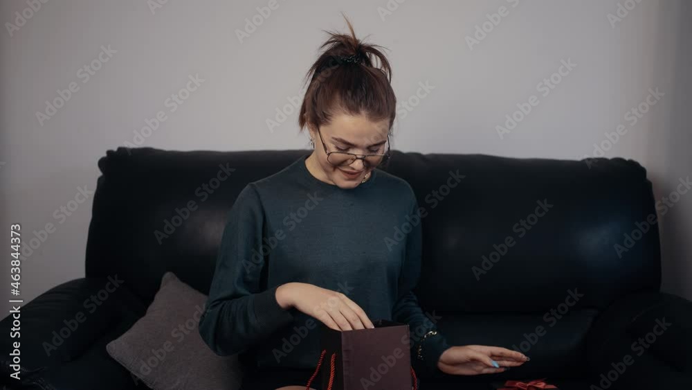 The young redhead, with glasses that opens the gift and surprised looks at its contents. Dressed in a dark green blouse, sitting on a black sofa, at home. The concept of the woman relaxing at home. 4k The young redhead, with glasses that opens the gift and surprised looks at its contents. Dressed in a dark green blouse, sitting on a black sofa, at home. The concept of the woman relaxing at home. 4k