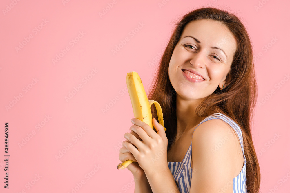 A young adult woman sexually holds a banana and laughs from embarrassment standing on a pink background A young adult woman sexually holds a banana and laughs from embarrassment standing on a pink background