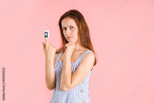 Portrait of a middle-aged woman in a device for measuring the level of oxygen in the blood. Pulse oximeter on the finger of a woman standing on a pink background