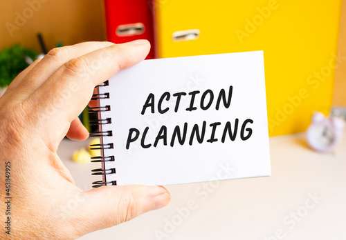 A male hand holds a notepad on a spring with the written ACTION PLANNING text. Office space.