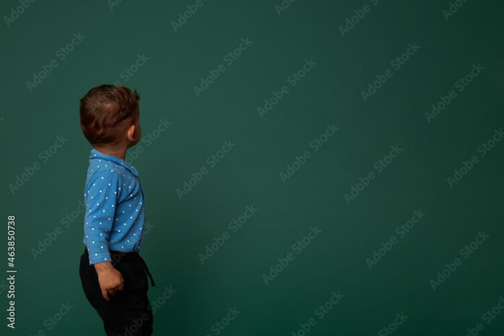 Back view picture of little boy with short haircut in blue shirt and black pants looking at green studio wall with copy space for your advertising content. Rear view. Childhood, curiosity Back view picture of little boy with short haircut in blue shirt and black pants looking at green studio wall with copy space for your advertising content. Rear view. Childhood, curiosity