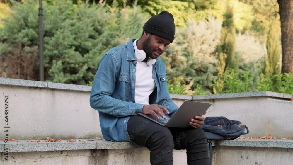 Positive African man in headphones texting by laptop on a concrete bench Positive African man in headphones texting by laptop on a concrete bench