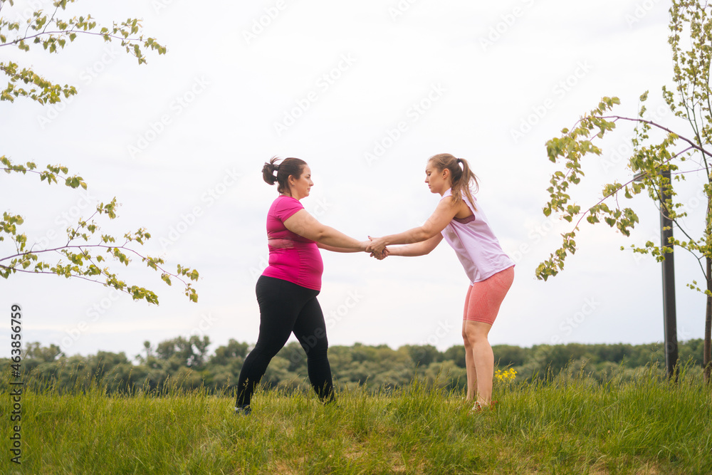 Wide shot of professional fitness female trainer giving personal training to overweight young woman outdoor in summer day. Instructor help fat woman lose weight outside doing squats outside. Wide shot of professional fitness female trainer giving personal training to overweight young woman outdoor in summer day. Instructor help fat woman lose weight outside doing squats outside.