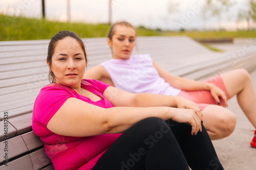 Close-up of tired obese young woman resting by bench after intense exercise with personal trainer outdoor in city park. Training from athletic lady for fat female with big abdomen wearing sportswear.