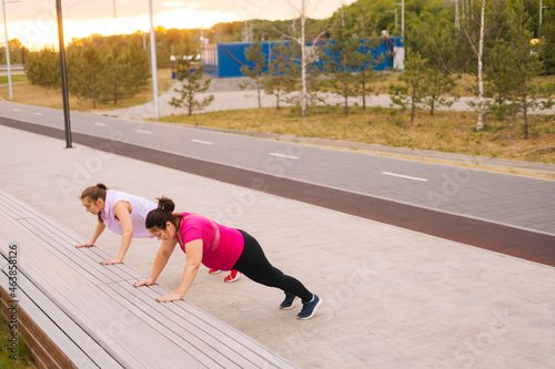 Wide shot of personal training from athletic lady for obese female with big abdomen outdoor in cloudy summer evening. Fat caucasian woman performing push up exercise with female trainer using bench.