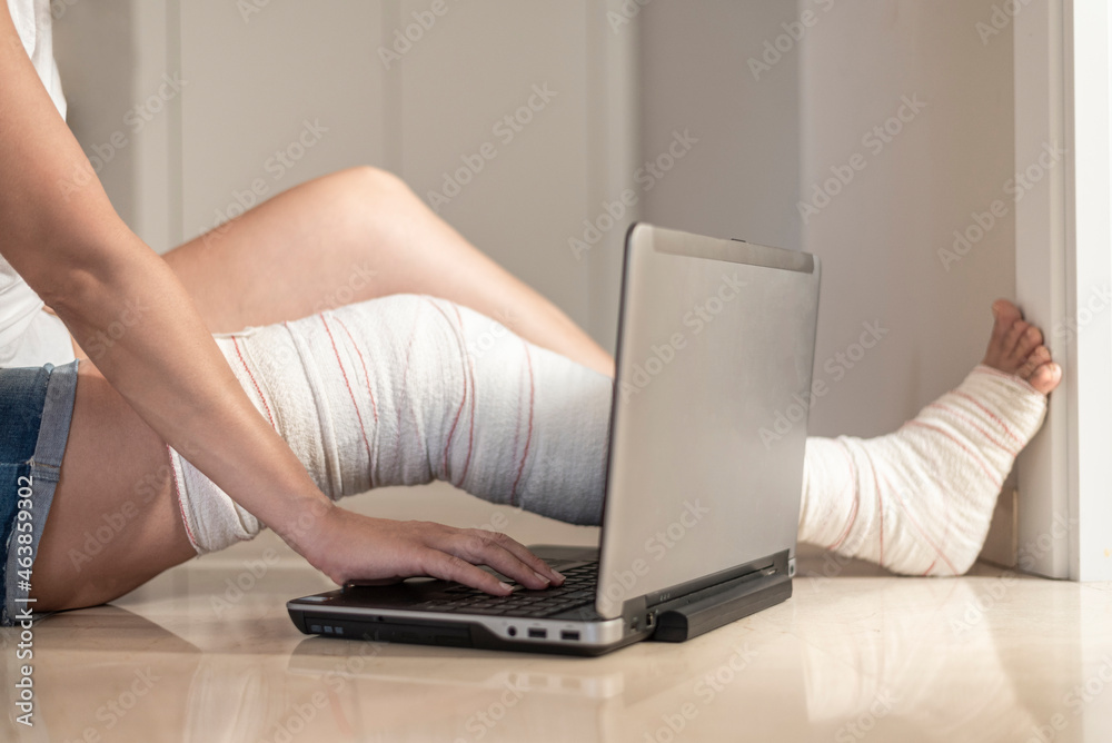 Fototapeta premium young woman sitting on the floor with bandaged leg typing on laptop