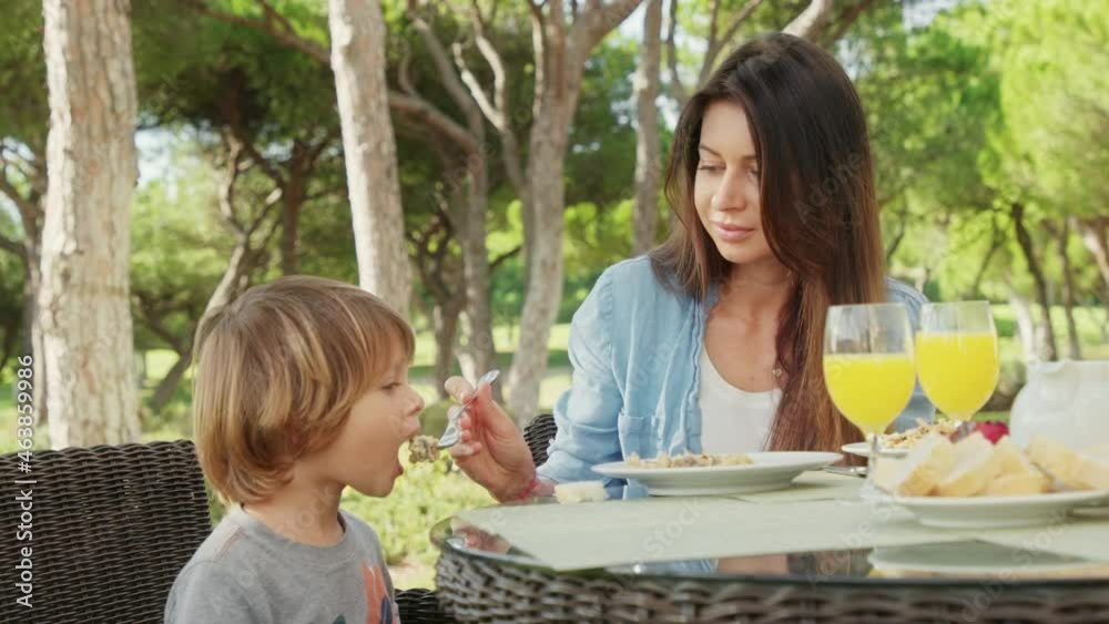 Cute boy enjoys his breakfast and fruity drink, while sitting by his mom. Beautiful caucasian mother feeds her sleepy son at the hotel terrace, surrounded with green garden. High quality 4k footage Cute boy enjoys his breakfast and fruity drink, while sitting by his mom. Beautiful caucasian mother feeds her sleepy son at the hotel terrace, surrounded with green garden. High quality 4k footage