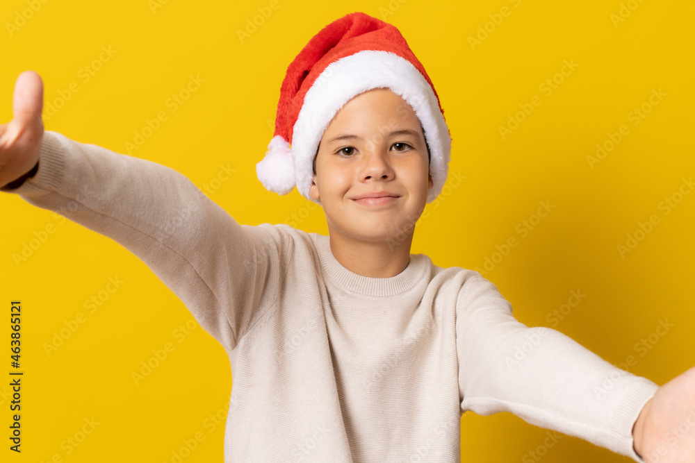 kid boy in santa hat against yellow wall looking at the camera smiling with open arms for hug. Cheerful expression embracing happiness. kid boy in santa hat against yellow wall looking at the camera smiling with open arms for hug. Cheerful expression embracing happiness.