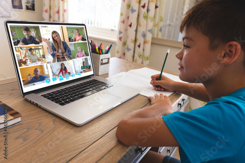 Caucasian boy using laptop for video call, with waving diverse elementary school pupils on screen