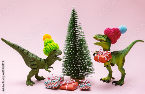 Funny plastic toy dinosaurs wearing knitted hat with  present boxes and evergreen tree on a pink background. © dvulikaia