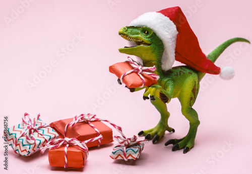 Funny plastic toy dinosaur wearing Santa Claus hat with  present boxes on a pink background.
