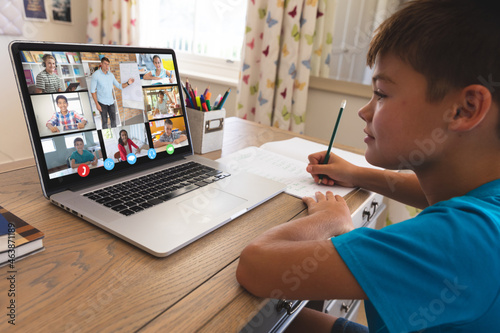 Smiling caucasian boy using laptop for video call, with diverse elementary school pupils on screen