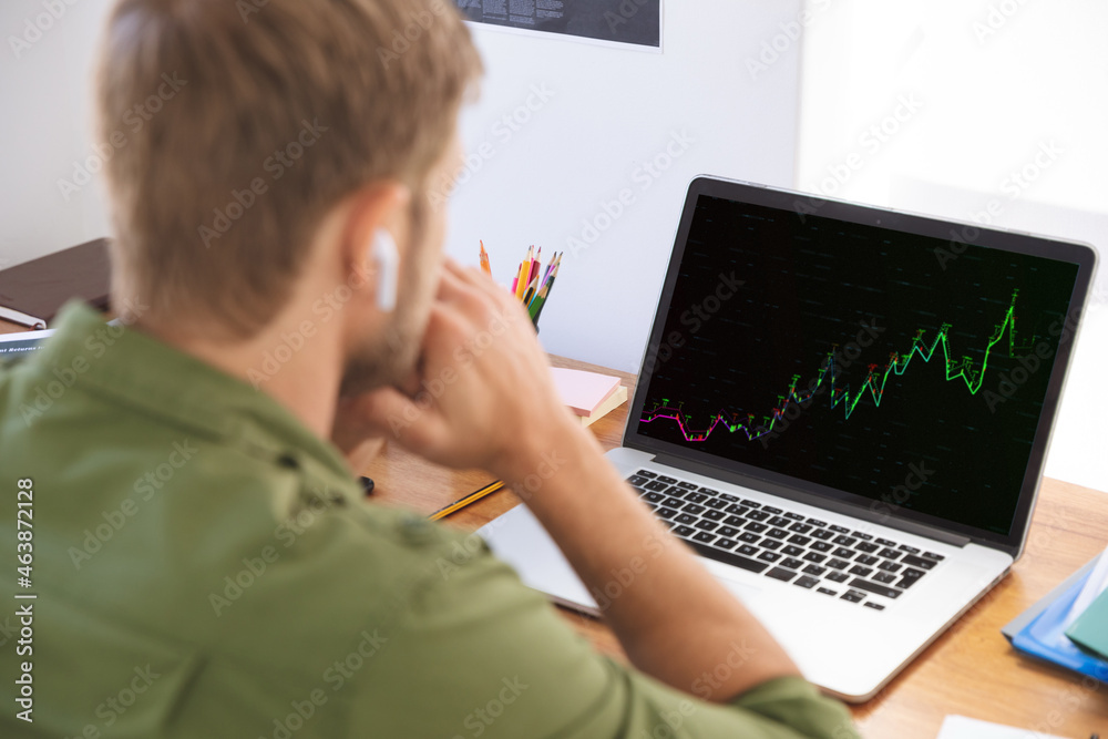 Caucasian businessman sitting at desk using laptop with graph and statistical data on screen Caucasian businessman sitting at desk using laptop with graph and statistical data on screen