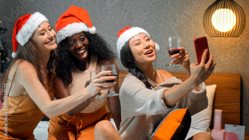 young girls European, Asian and African in red New Year's clothes and hats sit on a bed at a Christmas tree holding glasses of wine made photo on a phone, smiling and celebrating Christmas
