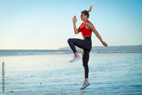 Attractive Strong Young Fit Athletic Blonde Pony Tail Woman Working Out Jumping on Beach Sand in Water Ocean Wearing Red Shirt Yoga Pants at Sunset with Blue Sky Background and Rocks