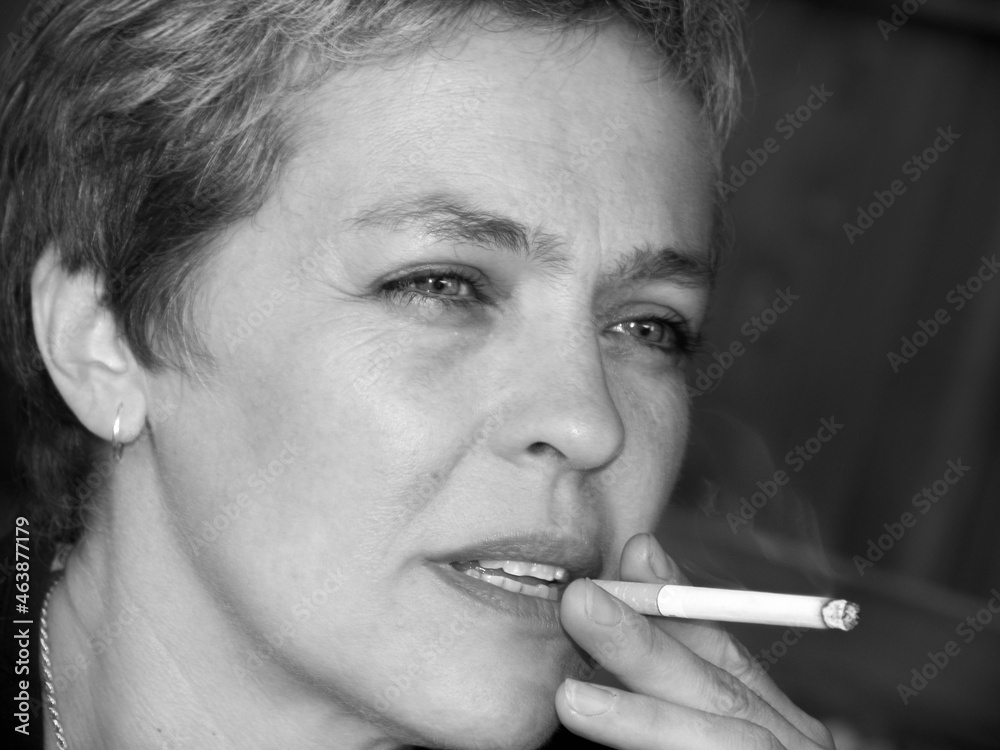 Obraz premium Dreaming middle age woman with the short hair cut, looking forward and thinking, dreaming, making puff on cigarette, holding the burnt cigarette with two fingers.