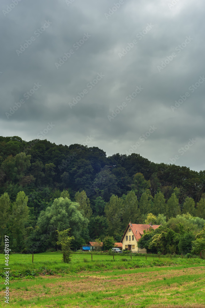 House with an off road vehicle on the edge of a hilly forest under a dark cloudy sky.