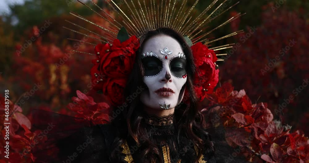 Portrait of young woman with sugar skull makeup and red roses dressed in black costume of death as Santa Muerte against background of autumn forest. Day of the Dead or Halloween concept.
