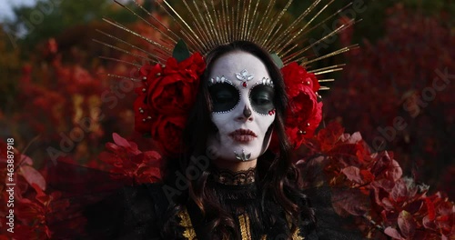 Portrait of young woman with sugar skull makeup and red roses dressed in black costume of death as Santa Muerte against background of autumn forest. Day of the Dead or Halloween concept.