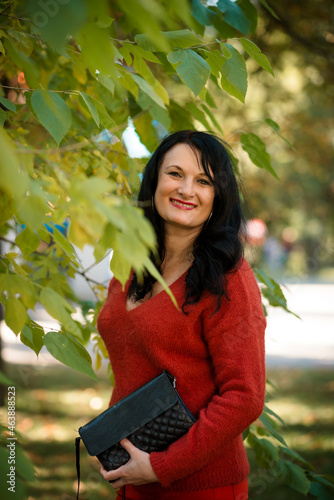 A woman with dark hair is smiling in the park near a green tree. In a red sweater.