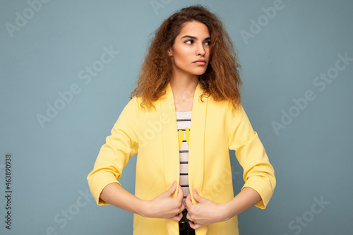 Young self-confident serious beautiful curly brunette woman with sincere emotions poising isolated over background wall with empty space wearing casual yellow jacket. Feelings concept