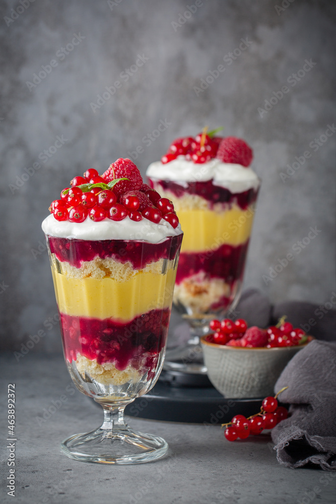 Trifles, layered dessert in glass with berry jelly, custard, sponge and whipped cream, decorated with fresh red currant and raspberry. Dark grey background, vertical image.