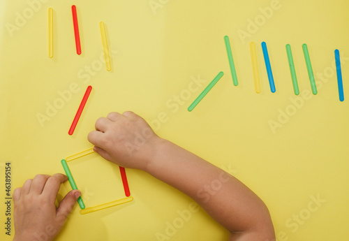 A small child collects a geometric figure from counting sticks. Children's hands with counting sticks isolated on a yellow background.