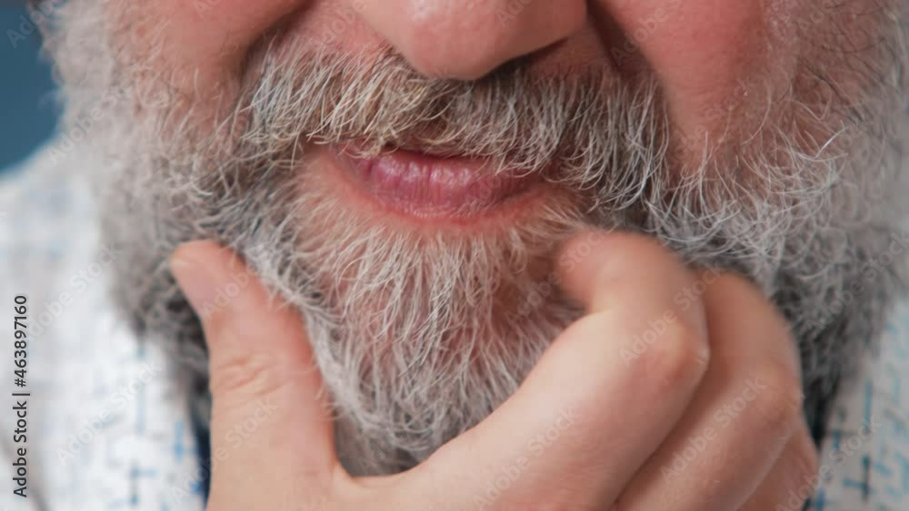 Close-up shooting. An adult man in a light shirt stands on a blue background and scratches his beard. There are characteristic emotions on his face. A bearded man. Close-up shooting. An adult man in a light shirt stands on a blue background and scratches his beard. There are characteristic emotions on his face. A bearded man.