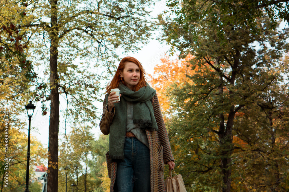 Stylish redhead young woman in coat with craft shopping bags walking in city park in fall Stylish redhead young woman in coat with craft shopping bags walking in city park in fall