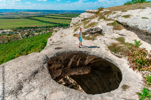 Obraz na plátně A young man on the edge of a precipice in the form of a huge hole in the ground among green fields and mountains on a sunny summer day