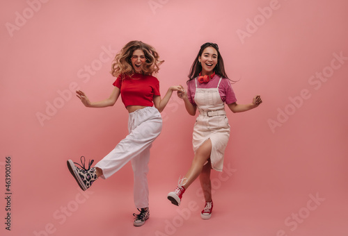 Full shot of cute european young hipsters dancing on pink background with place for text. Brunette and blonde girls in bright T-shirts hold hands and jump in place. Happy summer mood concept