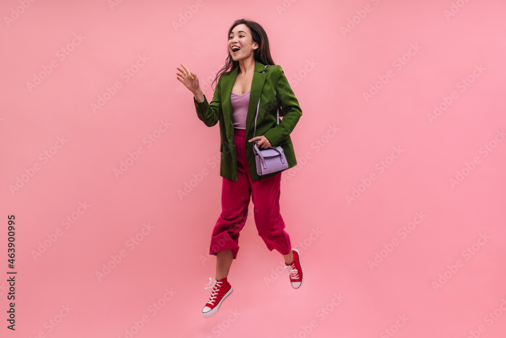 Full shot of beautiful whimsical asian woman greeting her best friends on street. Jumping and posing girl with white skin and straight black hair in green jacket, crimson pants.