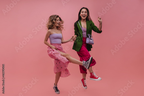 Attractive young women of asian and european appearance dancing and laughing in front of pink background. Models in bright spring outfits jump high and hold hands. Playful mood concept