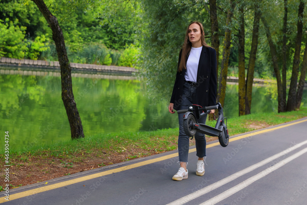 Fototapeta premium A young woman in stylish clothes walks along a bike path with a folded electric scooter