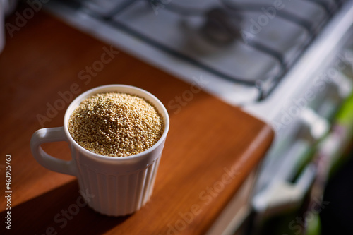 Organic fresh white pure selected wholesome quinoa grain seeds in ceramic mug on wooden countertop against kitchen stove background light of morning sun closeup