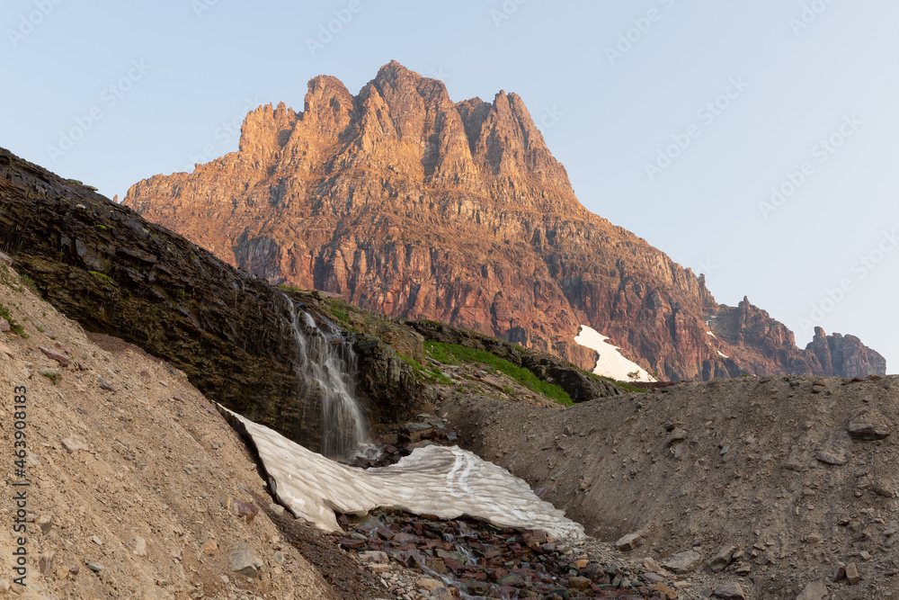 Clements Mountain catches the first of the morning's light as melting snow pours over a small waterfall towards the Hidden Lake trail in Glacier National Park Montana.	