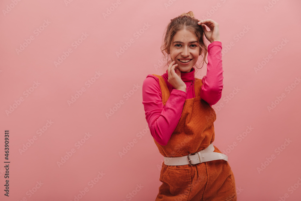 Medium shot of pretty young european blonde woman with joyful smile dancing against pink background. In orange jumpsuit with crimson long sleeve, her hands gently touch face. Medium shot of pretty young european blonde woman with joyful smile dancing against pink background. In orange jumpsuit with crimson long sleeve, her hands gently touch face.