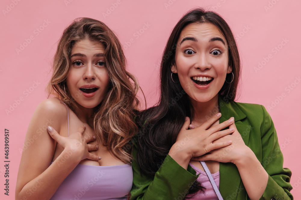 Close-up of sincerely happy asian and ukrainian women admire what they see on pink background. Blonde in lavender top with her brunette friend in green jacket. Human emotion and shock concept Close-up of sincerely happy asian and ukrainian women admire what they see on pink background. Blonde in lavender top with her brunette friend in green jacket. Human emotion and shock concept