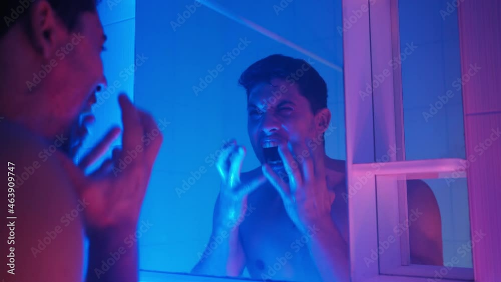 Angry man with stubble on his face washes his face in the sink and screams loudly in frustration and depression while looking in the mirror in neon lighting.