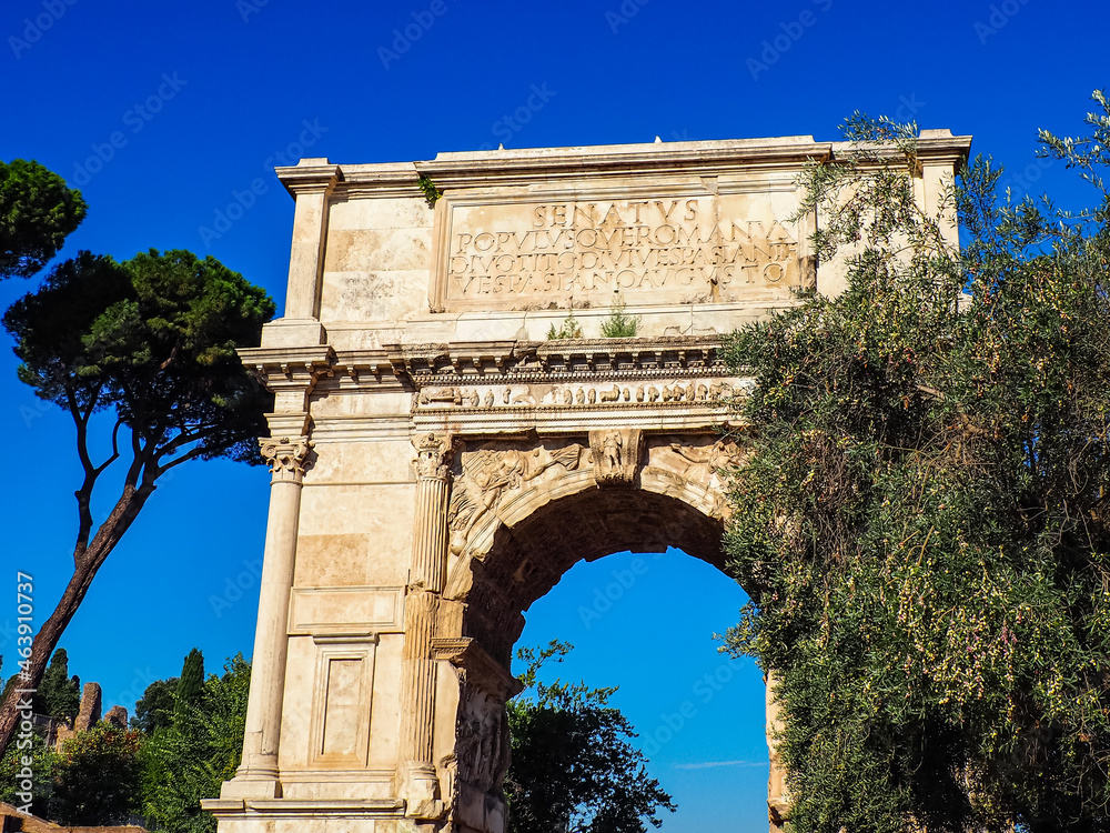 Arch of Titus, marble stone entrance to the Roman Forum, set against a blue sky with olive and pine trees and greenery, Forward facing  view