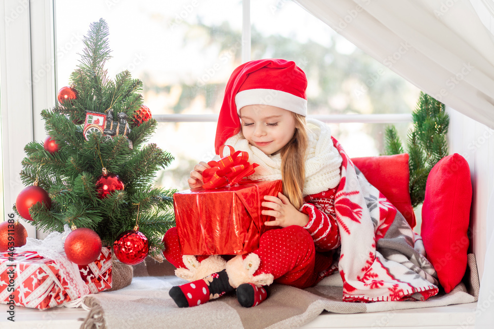 a cute girl child with a gift in her hands is sitting on the windowsill at the window of the house at the Christmas tree and waiting for the new year or Christmas in a red Santa Claus hat a cute girl child with a gift in her hands is sitting on the windowsill at the window of the house at the Christmas tree and waiting for the new year or Christmas in a red Santa Claus hat