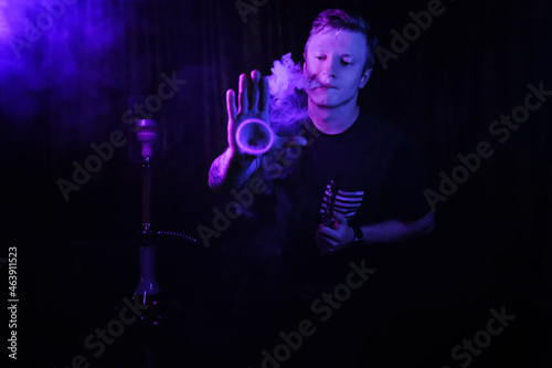 young guy smokes a hookah. neon light. emits smoke in the form of a ring.