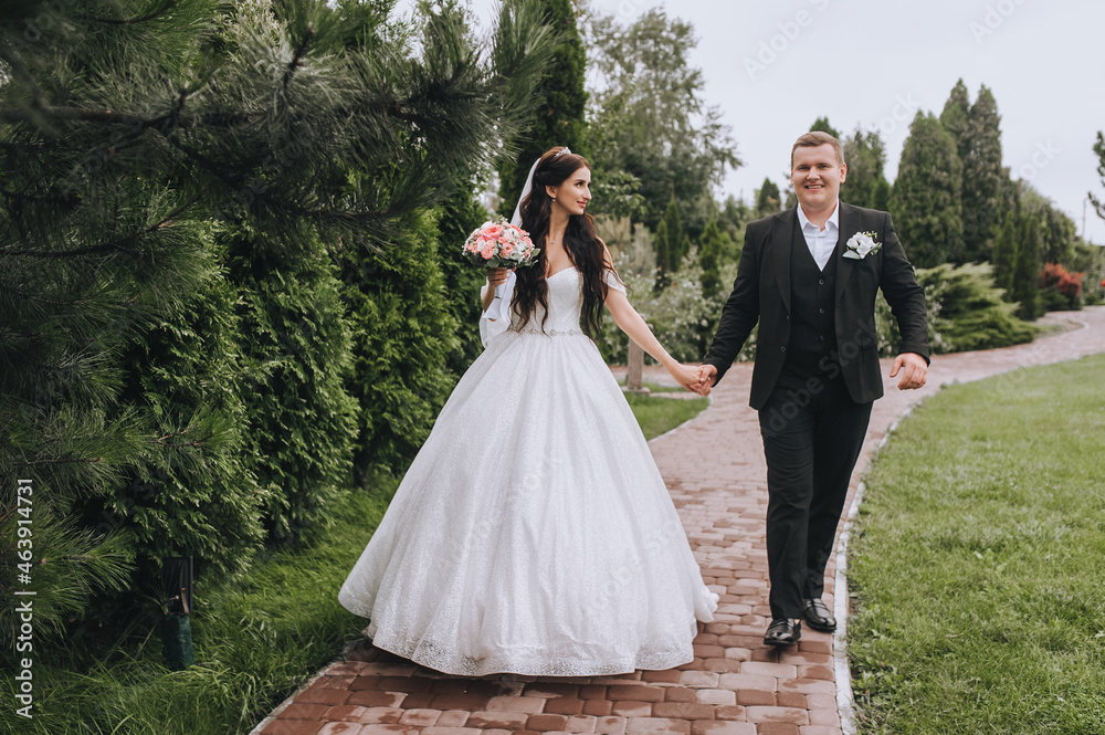 Fashionable groom in a black suit and a beautiful brunette bride in a white dress with a bouquet are walking along the park, road, holding hands. Wedding portrait of smiling newlyweds.