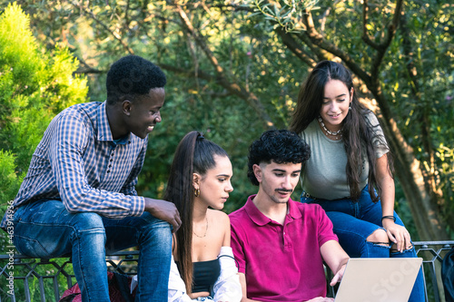 Four multiethnic friends looking at a laptop while sitting on a bench of a park