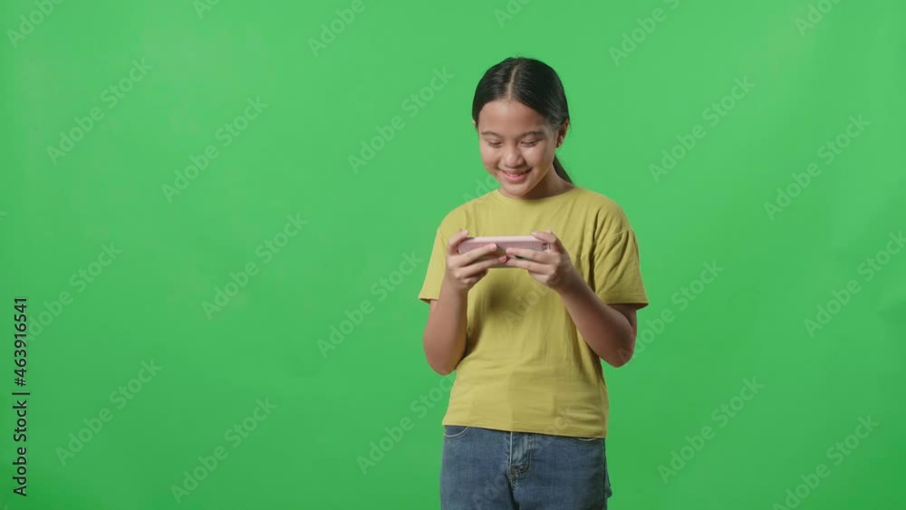 Smiling Young Asian Kid Girl Looking At The Phone Screen And Holding It In Her Hands While Standing On Green Screen In The Studio
Smiling Young Asian Kid Girl Looking At The Phone Screen And Holding It In Her Hands While Standing On Green Screen In The Studio