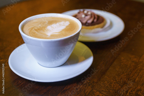 cup of cappuccino with Latte art on top is on the wooden table and cake in soft focus on background