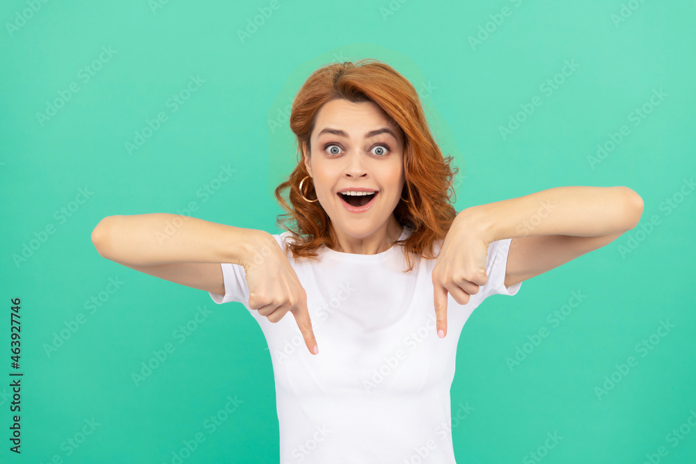 amazed redhead woman pointing finger down on blue background, copy space, shopping sales. amazed redhead woman pointing finger down on blue background, copy space, shopping sales.